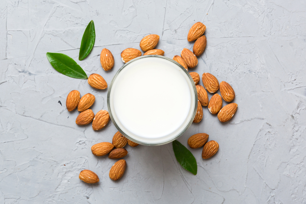 Yukarıdan çekilmiş beyaz zeminin üzerinde 1 bardak badem sütü, bardağın etrafı bademlerle çevrelenmiş.